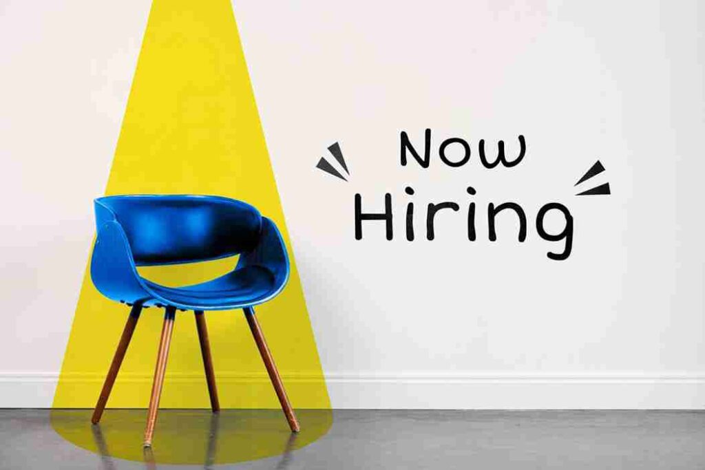 A modern blue chair with wooden legs against a white wall, featuring a yellow spotlight effect and "Now Hiring" text.